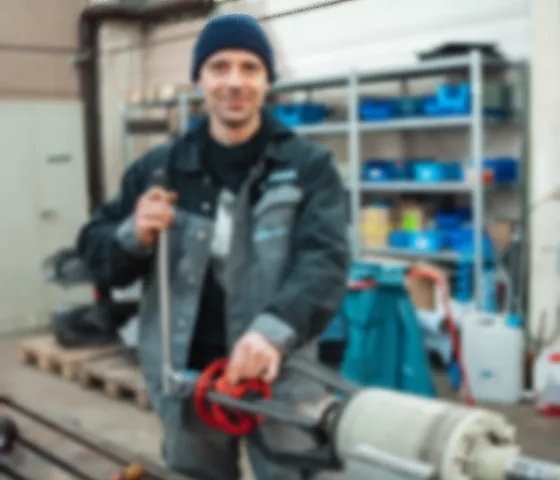 Mann in Arbeitskleidung und Mütze hält Werkzeug in einer Werkstatt mit Regalen im Hintergrund.