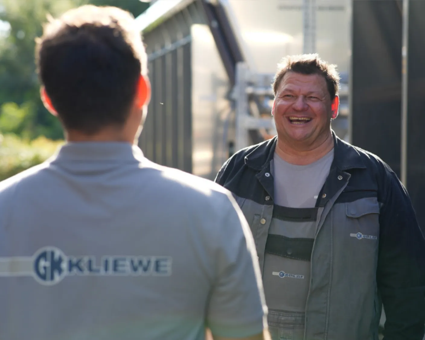 Zwei Männer in Arbeitskleidung mit dem Logo KLIEWE, ein Mann lächelt, während sie sich im Freien unterhalten.
