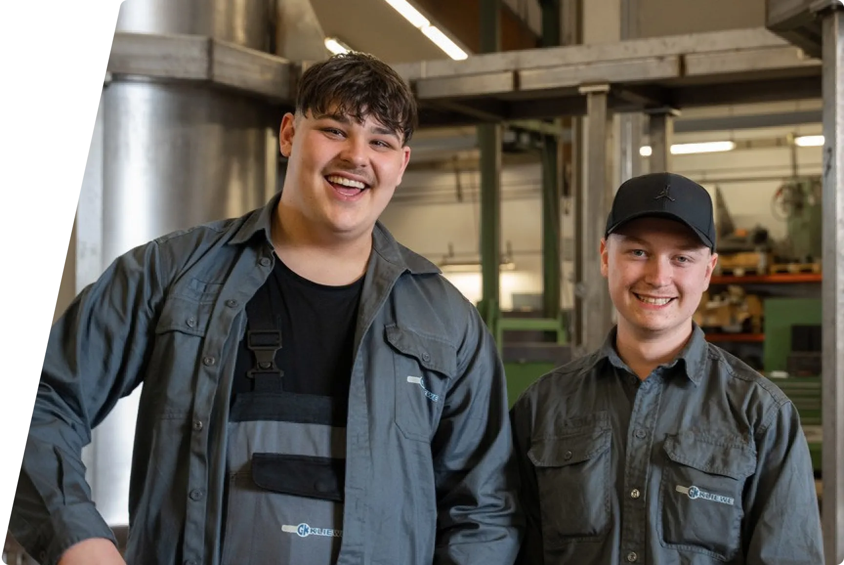 Zwei junge männliche Arbeiter in grauer Arbeitskleidung lächeln in einer Werkstatt.