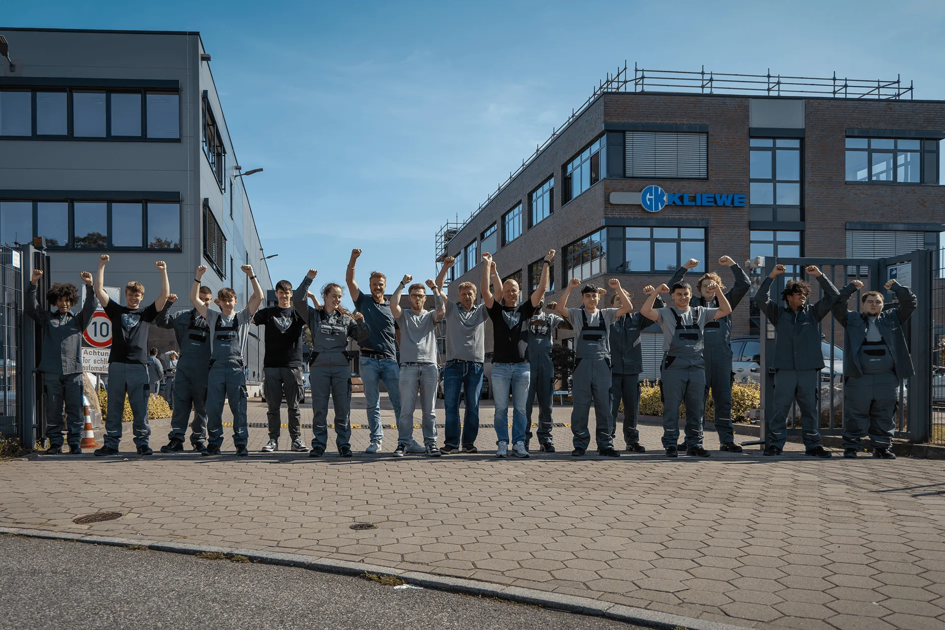 Gruppenfoto von Mitarbeitern vor einem Gebäude mit dem Logo Kliewe, die mit erhobenen Fäusten jubeln.