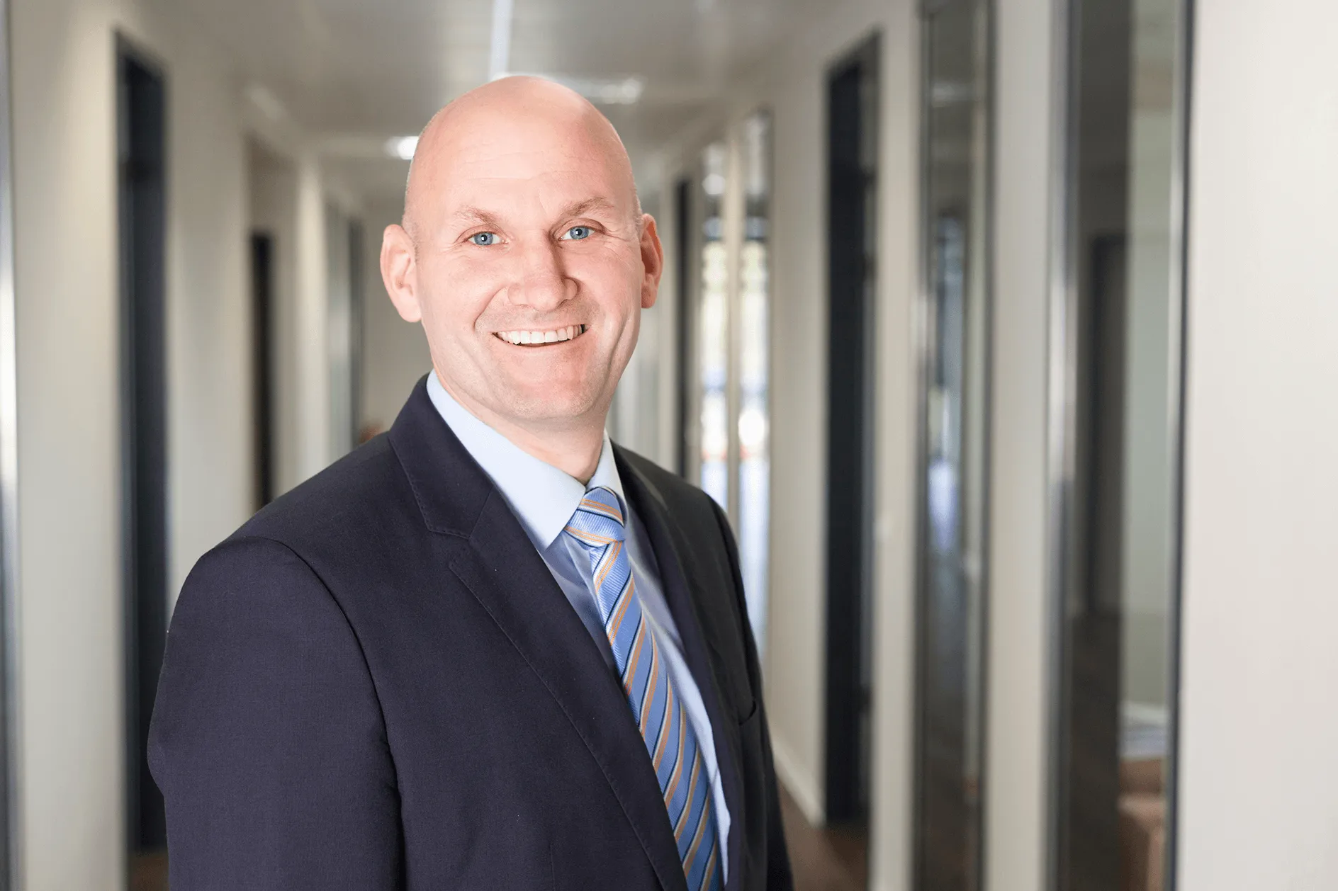 Lächelnder glatzköpfiger Mann in einem dunklen Anzug und gestreifter Krawatte steht in einem Büroflur.