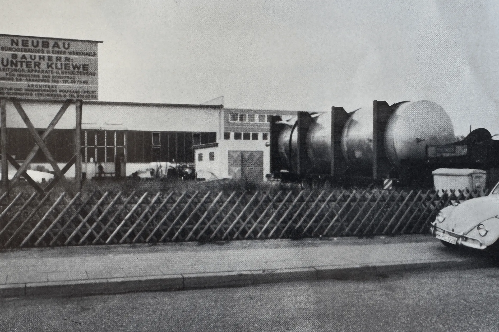 Schwarz-weiß-Foto eines Fabrikgebäudes mit einem großen Zylindertank auf einem Anhänger und einem Auto am Straßenrand.