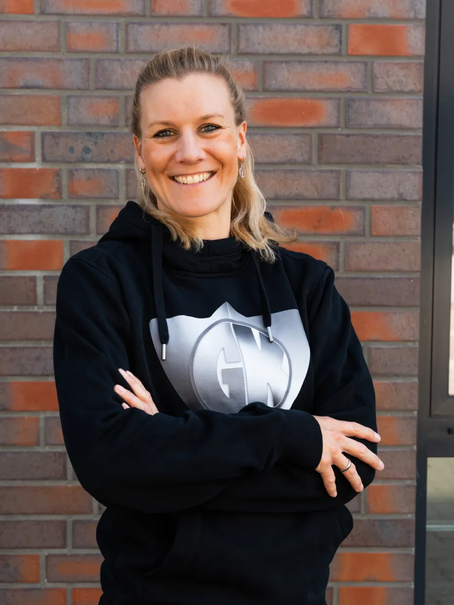Lächelnde Frau mit blonden Haaren, die einen schwarzen Kapuzenpullover mit einem silbernen GN-Logo vor einer Ziegelwand trägt.