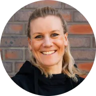 Fröhliche blonde Frau mit zusammengebundenem Haar vor einer Ziegelwand.