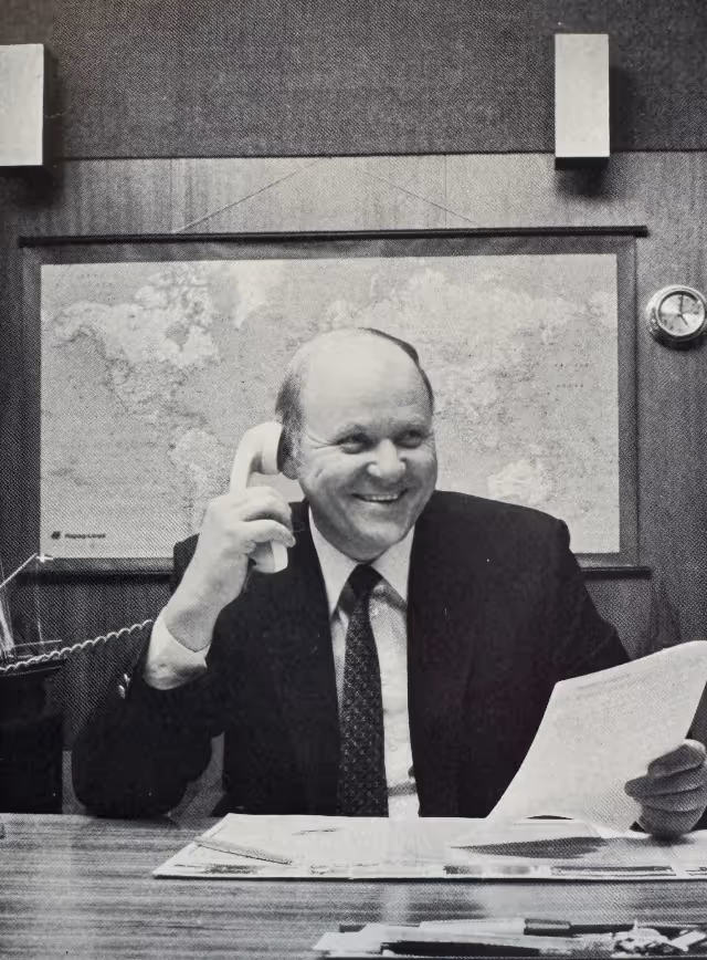 Geschäftsmann in Anzug, der am Schreibtisch sitzt, lächelnd ein Telefon hält und Papiere betrachtet, mit Weltkarte und Uhr an der Wand im Hintergrund.