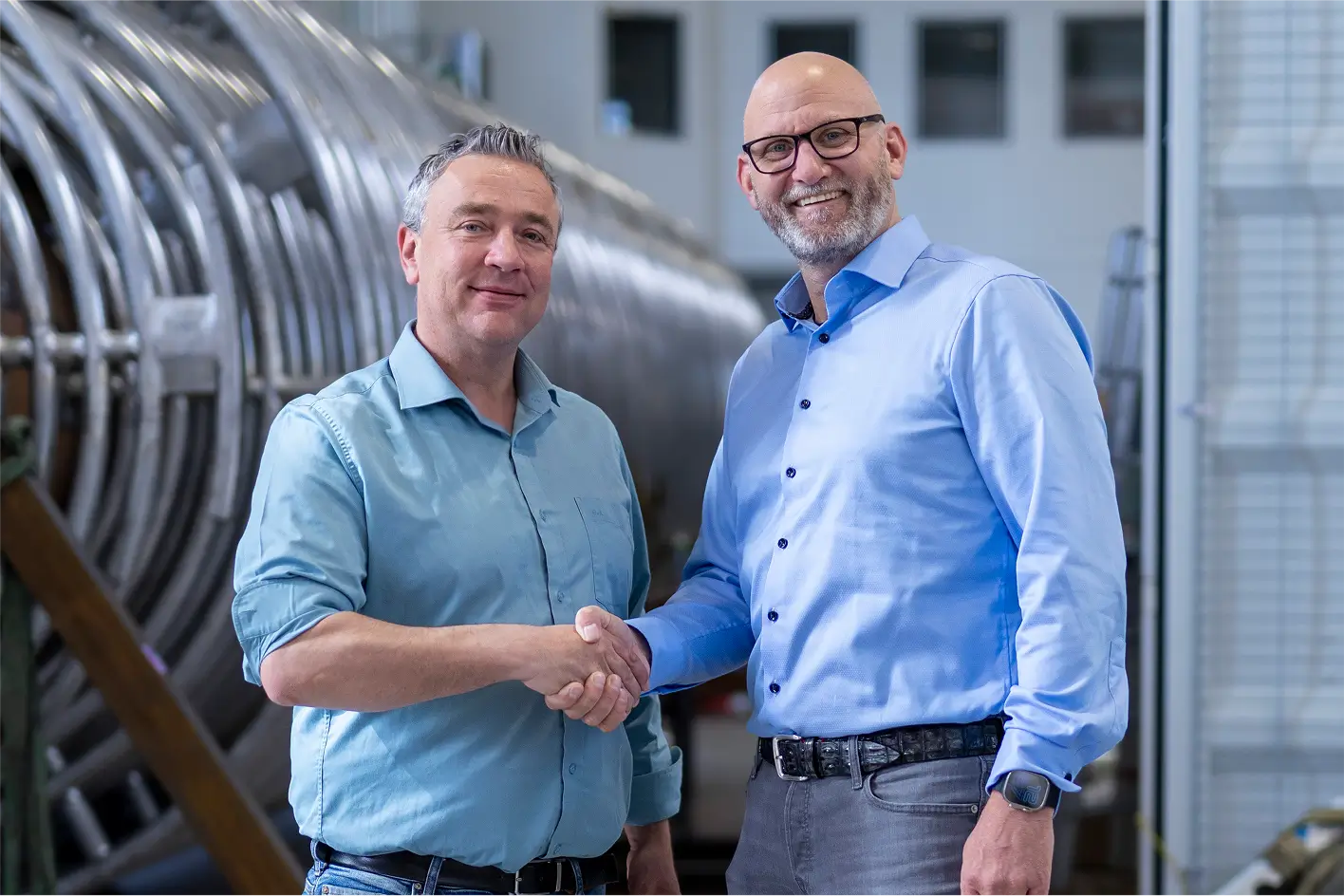 Zwei Männer in hellblauen Hemden schütteln sich in einer industriellen Umgebung die Hände und lächeln.