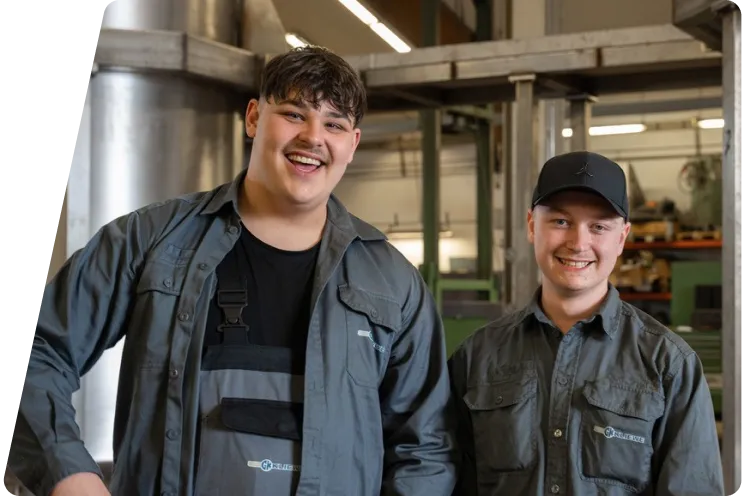 Zwei junge Männer in Arbeitskleidung lächeln in einer industriellen Werkstatt.