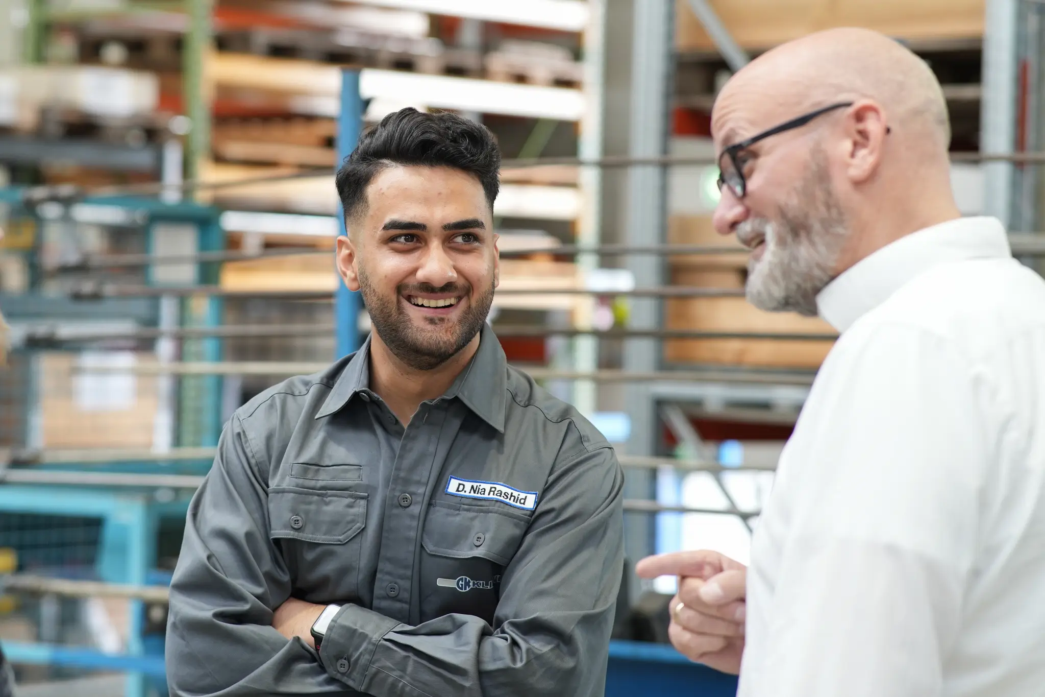 Zwei Männer unterhalten sich freundlich in einer industriellen Werkstattumgebung, einer trägt graue Arbeitskleidung.