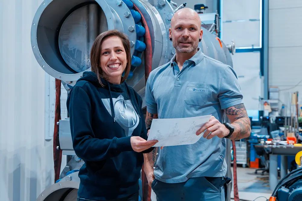 Zwei Personen stehen in einer Werkstatt vor großer Industriemaschine, halten technische Zeichnungen und lächeln in die Kamera.