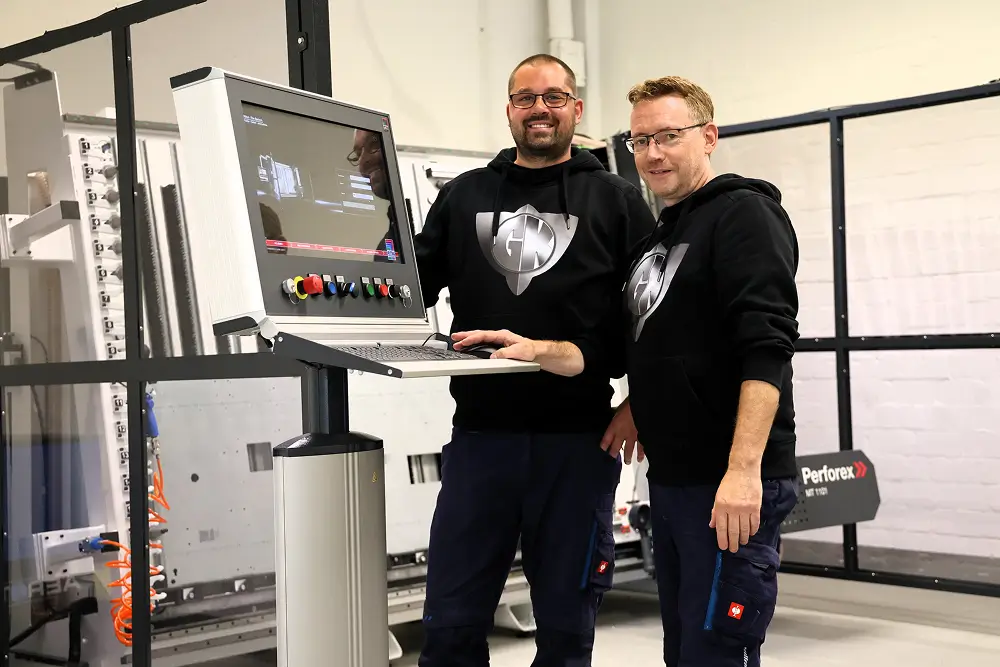 Zwei Männer in schwarzen Kapuzenpullovern bedienen gemeinsam eine industrielle Steuerung mit Bildschirm in einer Werkstatt.