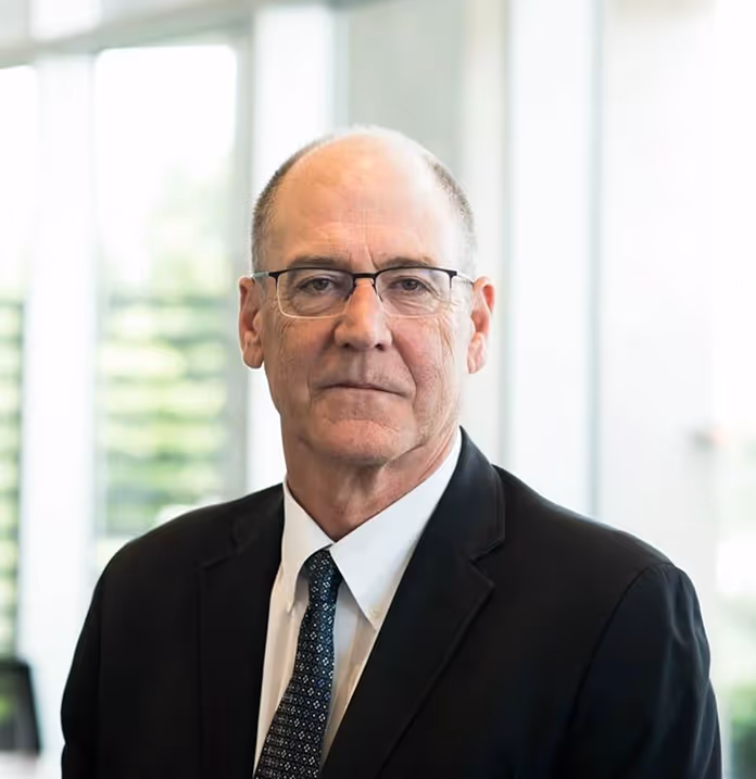 Portrait of a middle-aged man wearing glasses, a dark suit, white shirt, and patterned tie in a bright office setting.