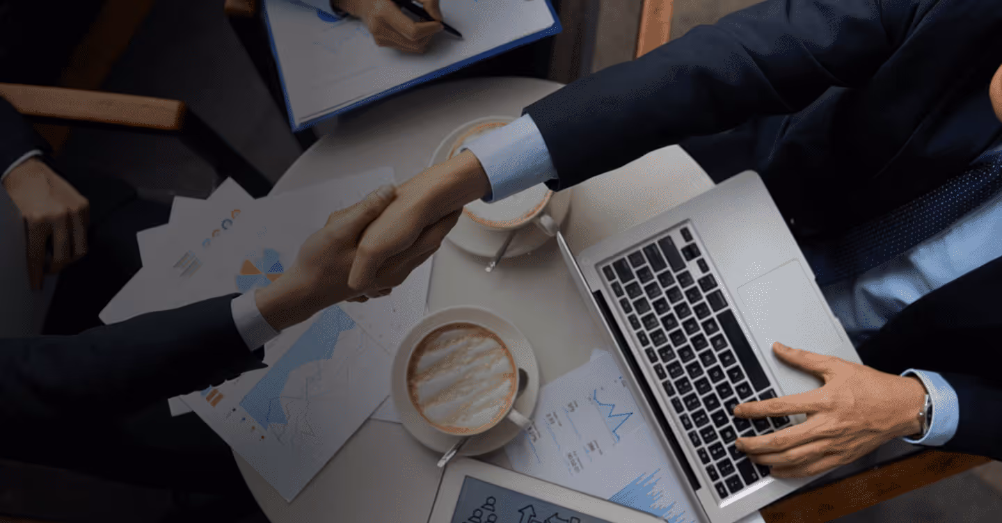 Two business professionals shaking hands over a table with a laptop, coffee cups, and financial charts.