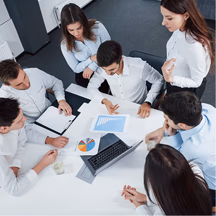 Six colleagues in formal shirts gathered around a table reviewing charts and a laptop during a meeting.