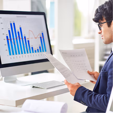 Person reviewing documents while sitting at a desk with a computer screen displaying a blue and red bar and line chart.