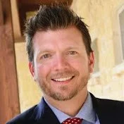 Smiling man with short dark hair and beard wearing a dark suit and red patterned tie.