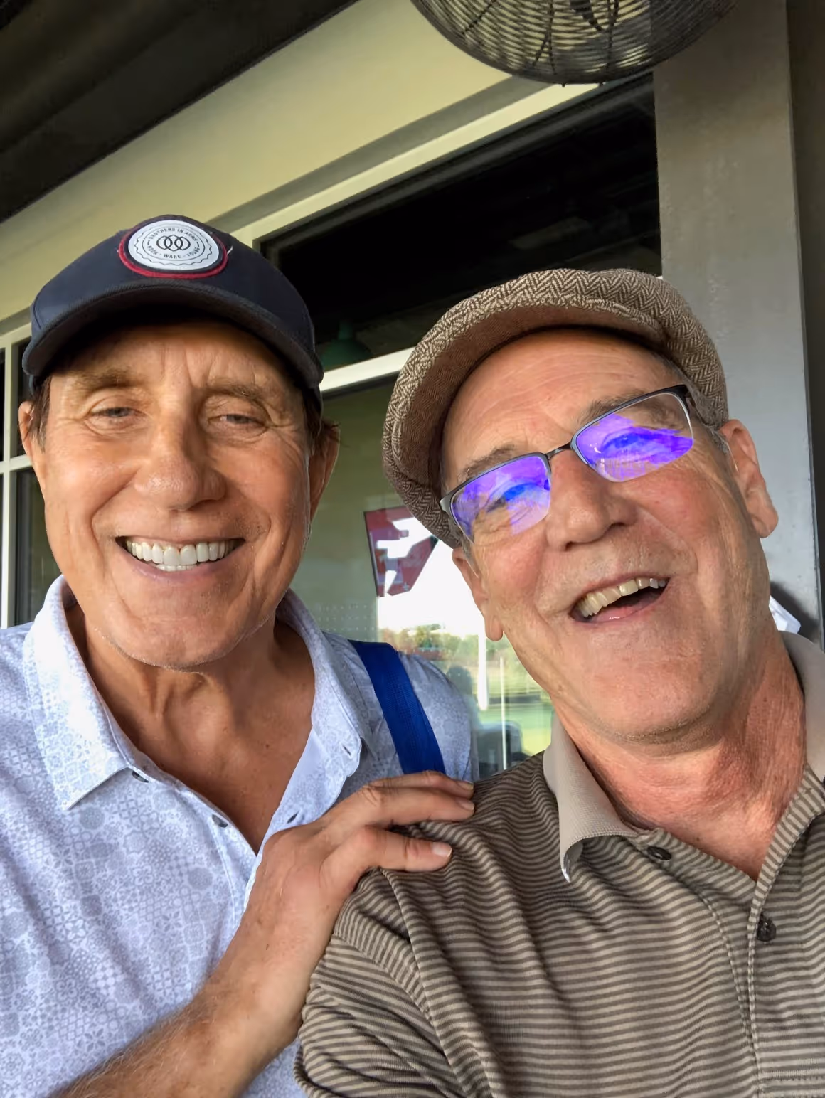 Two older men smiling together, one wearing a dark cap and white patterned shirt, the other wearing glasses and a brown striped polo shirt.