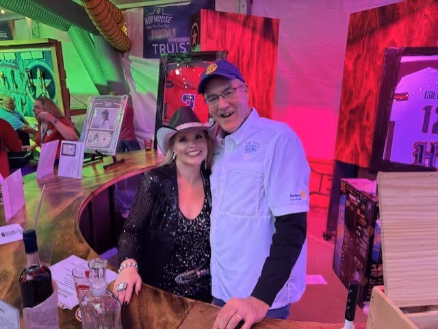 Smiling man and woman posing together behind a wooden bar counter in a colorful indoor setting.