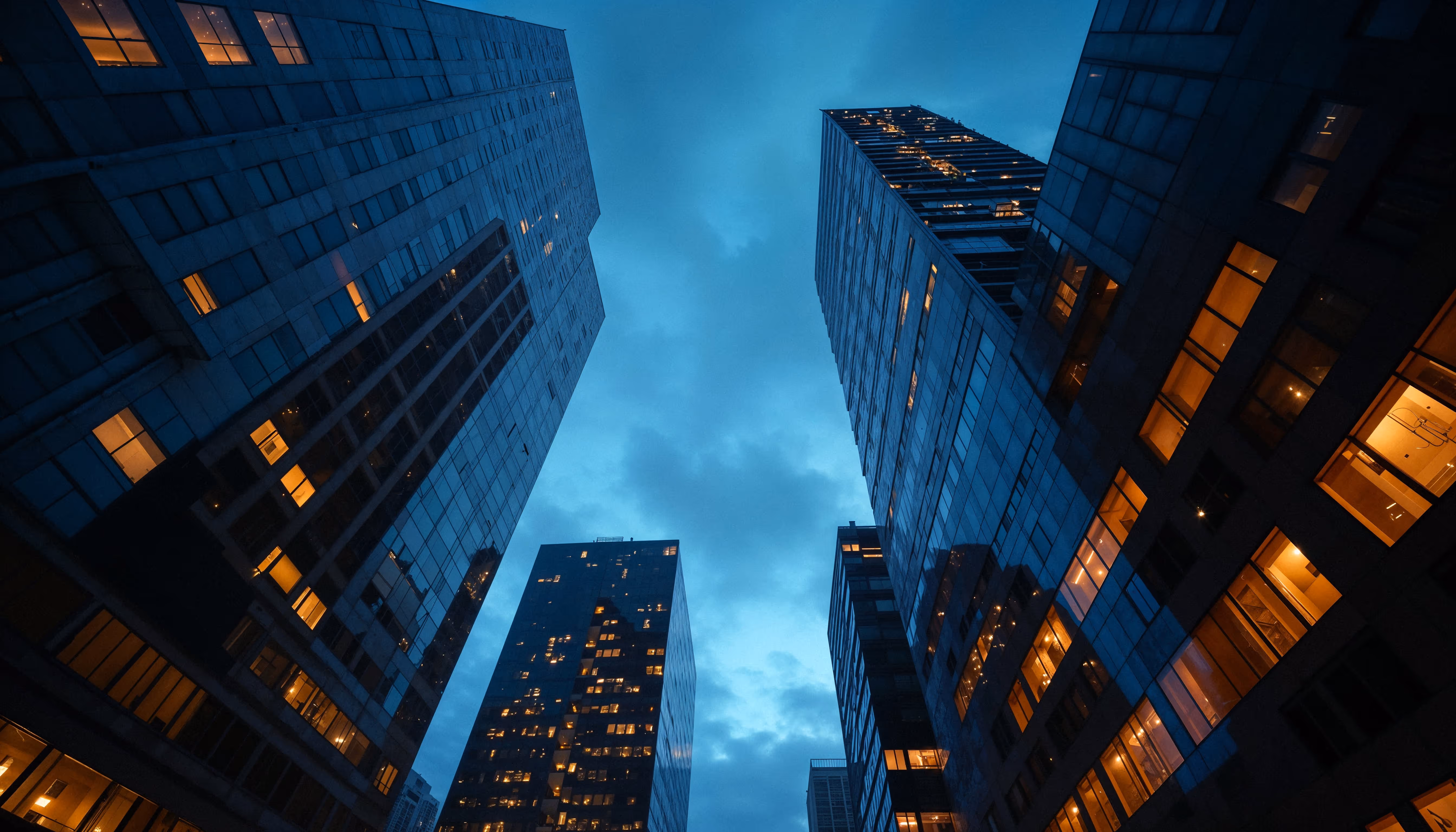 Looking up at tall skyscrapers with illuminated windows against a blue evening sky.
