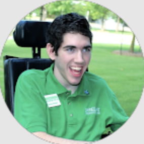 Smiling young man with dark hair sitting in a wheelchair outdoors wearing a green polo shirt.