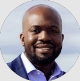 Smiling bald man with a beard wearing a blue blazer and light blue shirt against a light sky background.