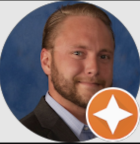Headshot of a man with light brown hair and beard, wearing a suit jacket, blue shirt, smiling against a blue background with an orange and white star badge overlay.