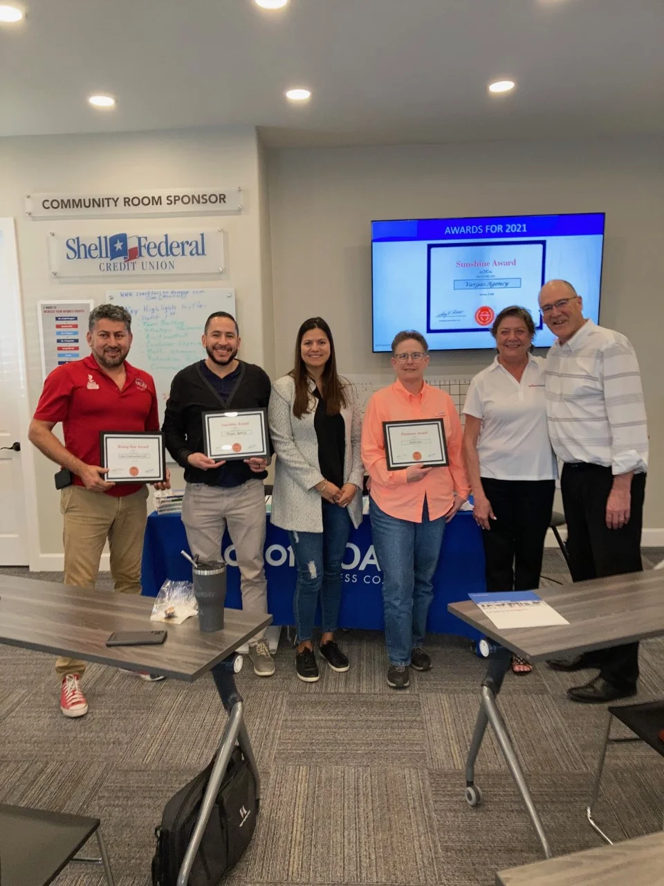 Six people standing indoors with three holding award certificates in front of a Community Room Sponsor sign and a screen showing awards for 2021.