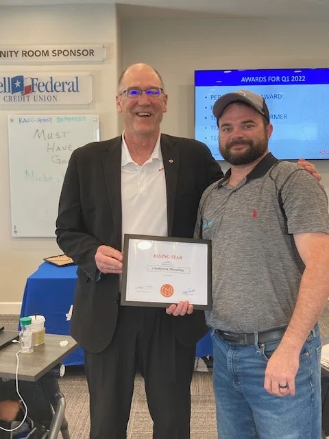 Two men smiling, one in a suit holding a 'Rising Star' award certificate and the other casually dressed with a cap, standing in an office setting.