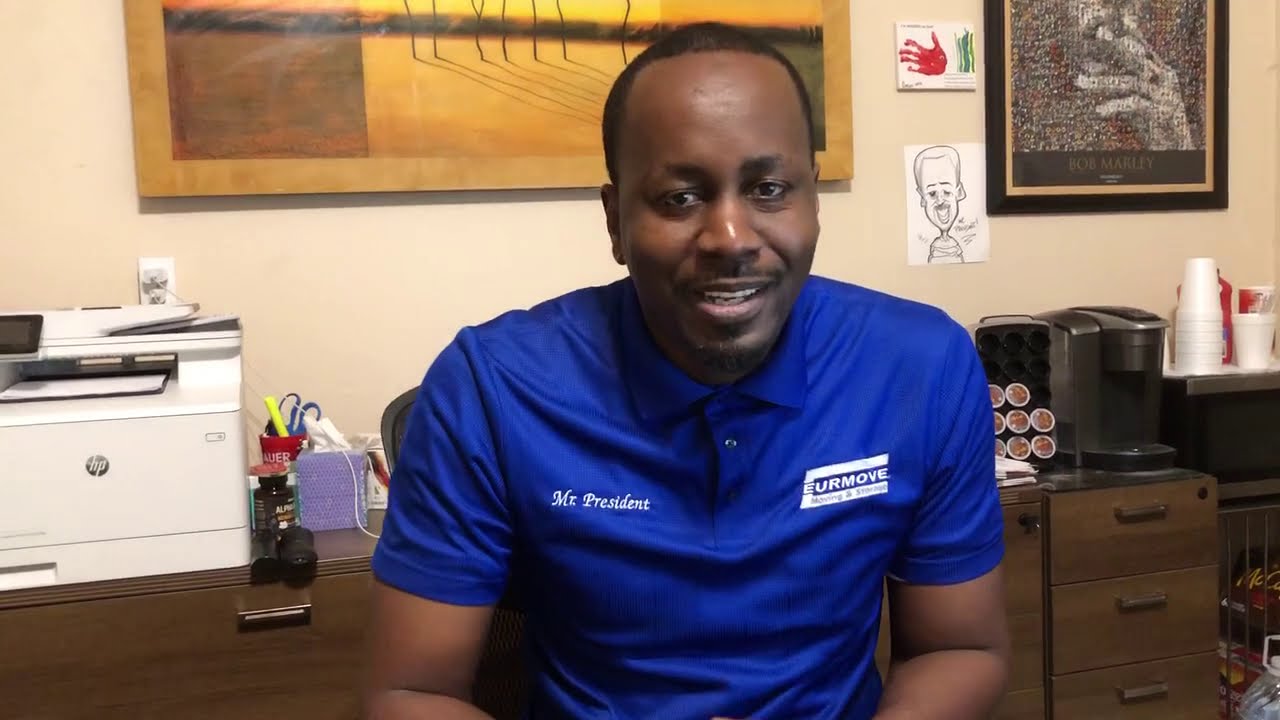 Man wearing a blue polo shirt with 'Mr. President' and 'EURMOVE Moving & Storage' logos, sitting in an office with artwork and office equipment behind him.