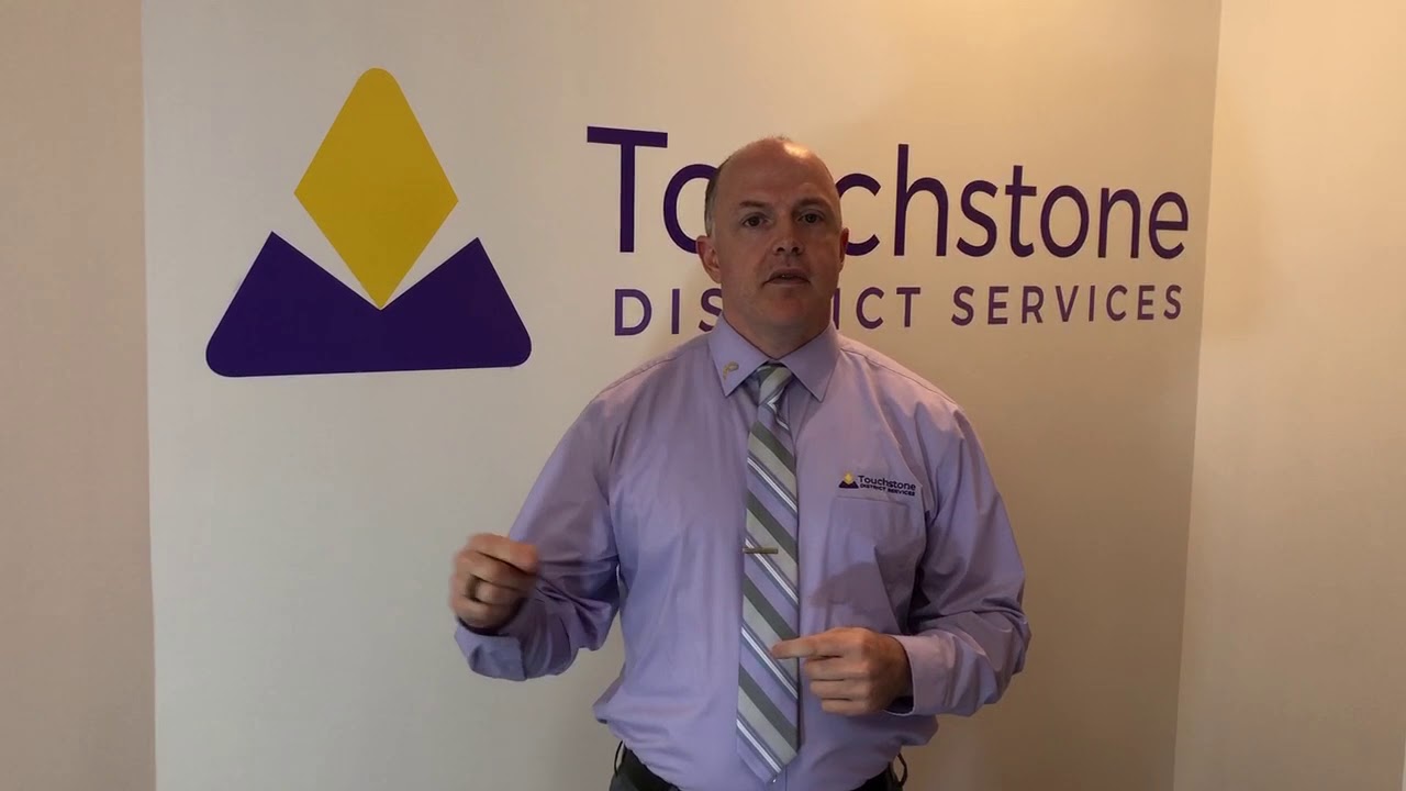 Man in purple shirt and striped tie standing in front of a wall with Touchstone District Services logo and text.