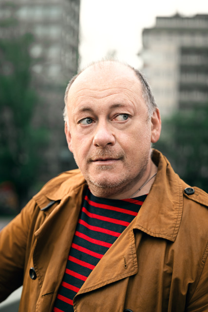 Ein Mann Ende 50 mit grauer Halbglatze und Dreitagebart, rotblau gestreiftem Shirt und einem braunen Trenchcoat