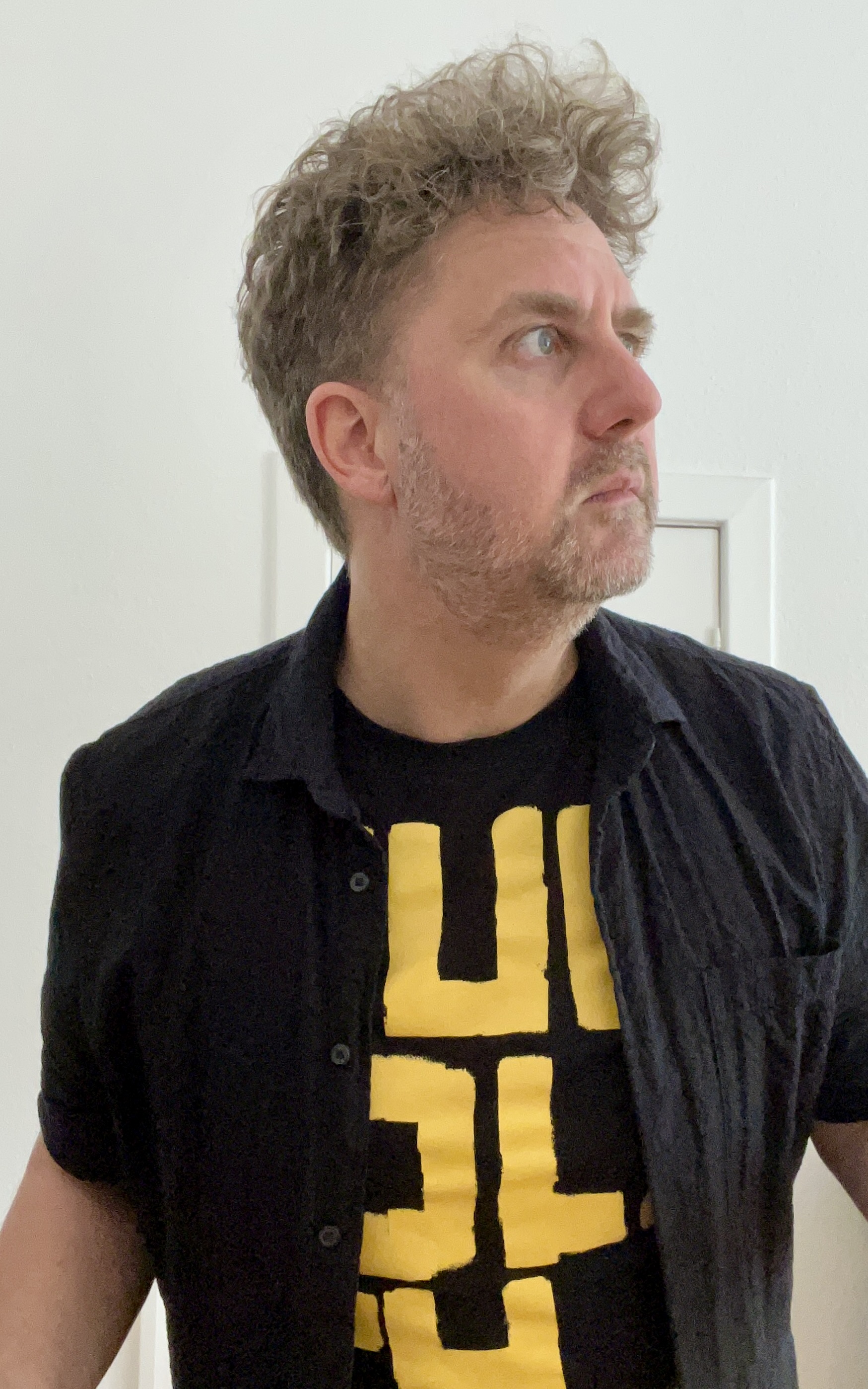 Ein Mann mit lockigen hellbraunen Haaren und Dreitagebart, er trägt ein schwarzgelbes T-Shirt und darüber ein offenes schwarzes Hemd.