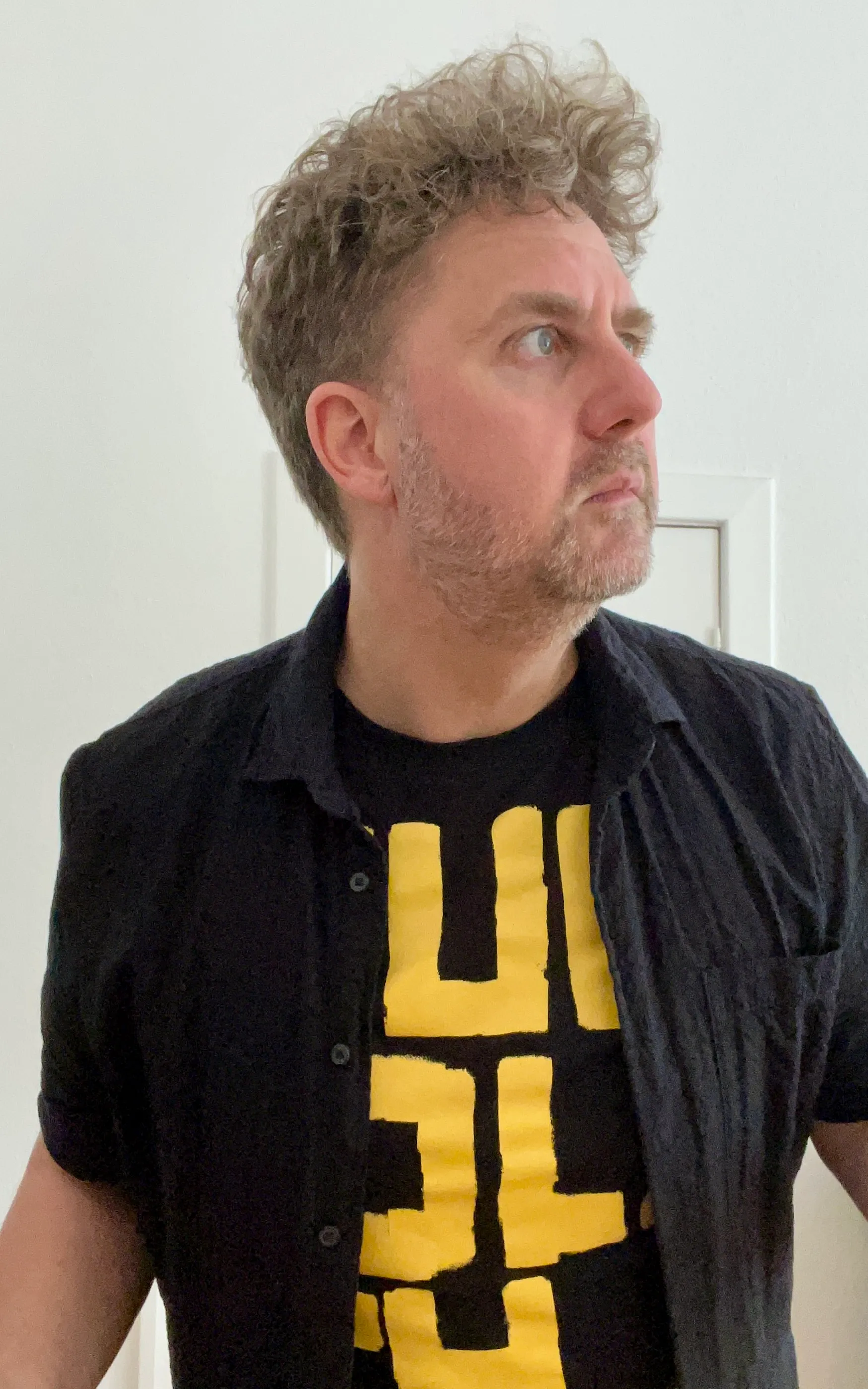 Ein Mann mit lockigen hellbraunen Haaren und Dreitagebart, er trägt ein schwarzgelbes T-Shirt und darüber ein offenes schwarzes Hemd.