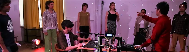 Zwei Personen an Mischpulten, um sie herum stehen sechs weitere Menschen im Halbkreis aufgestellt