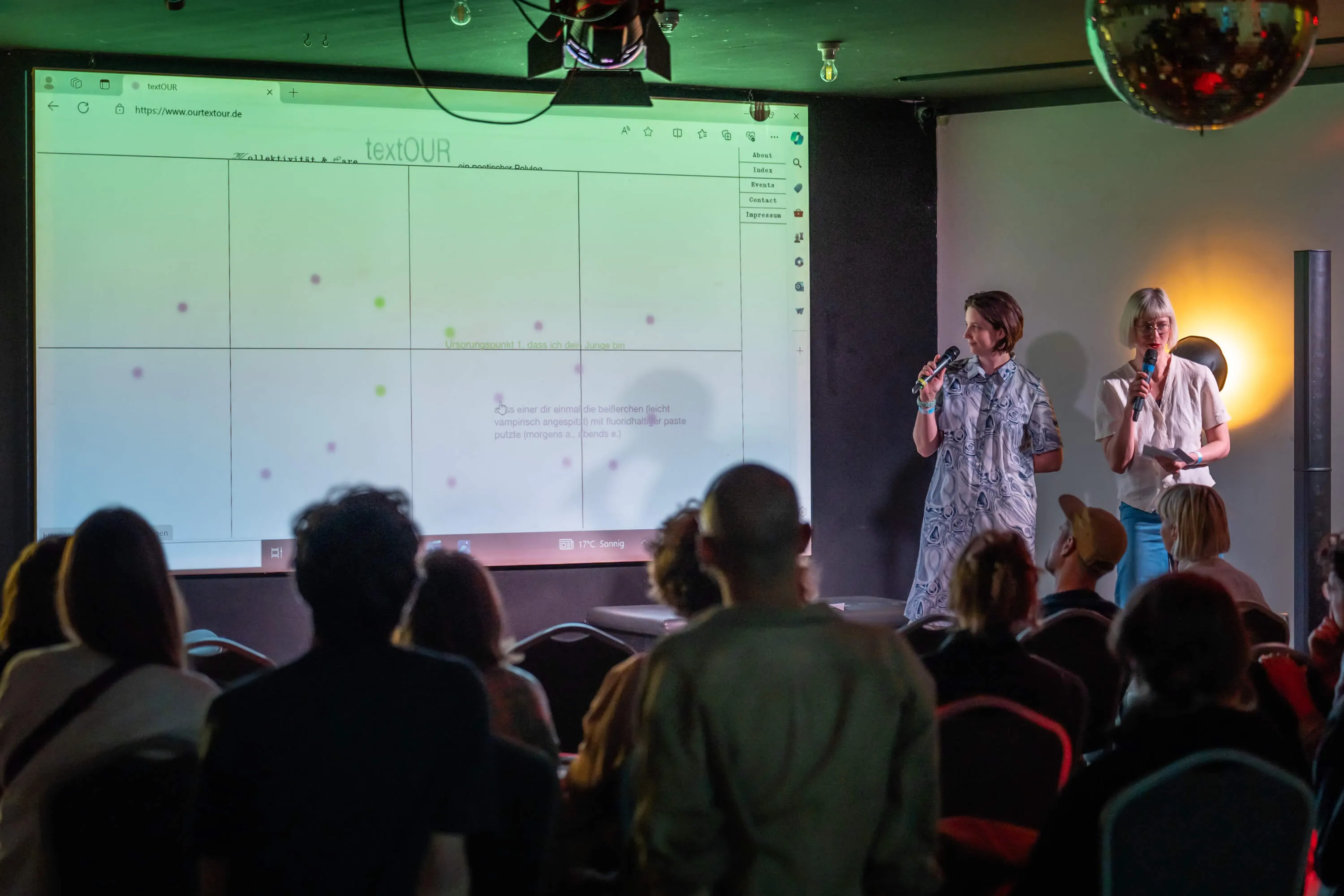 Zwei Mitglieder von das ad hoc stehen mit Mikrophonen neben einer Leinwand, auf die die textOUR-Website projiziert ist.