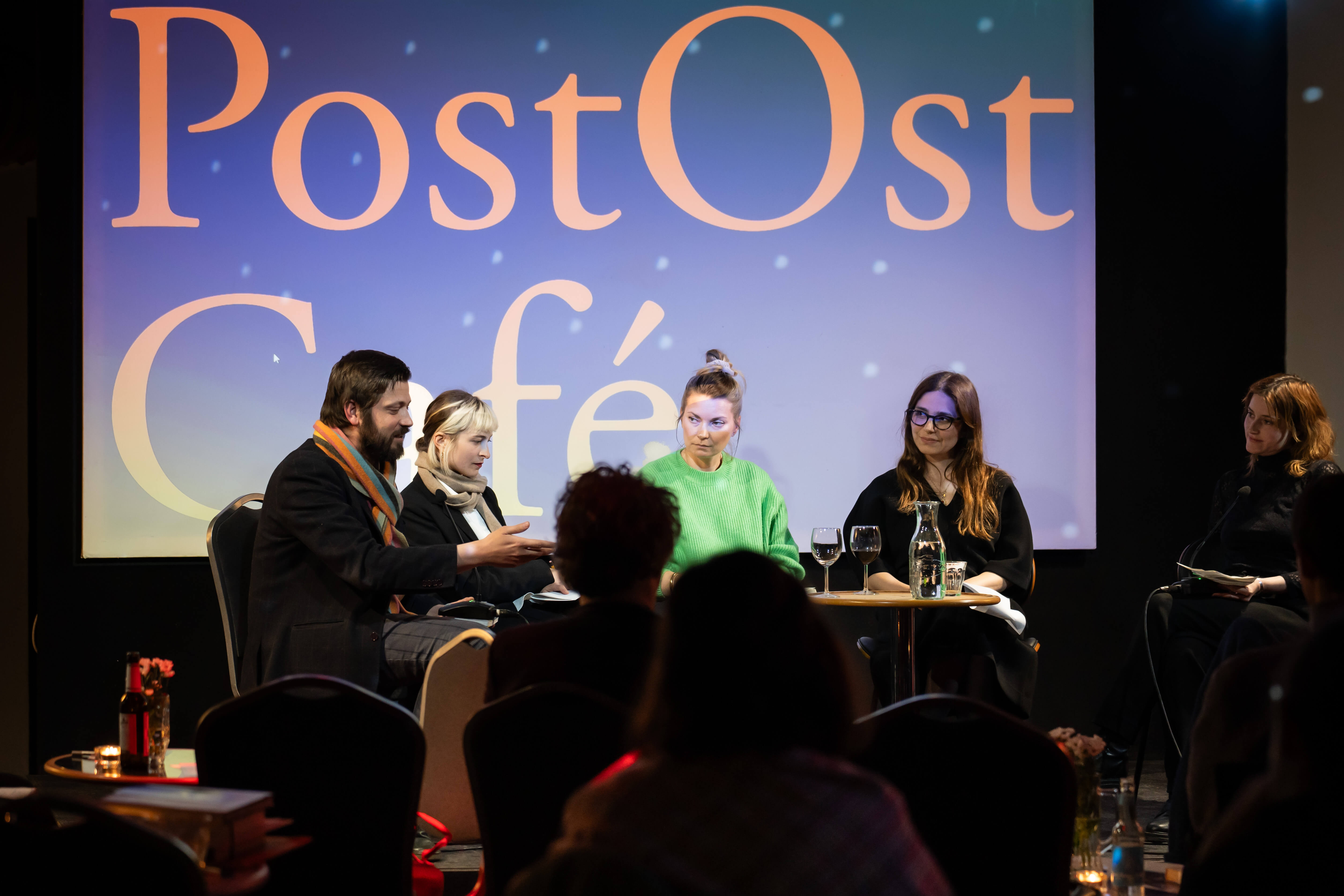 Fünf Menschen sitzen auf der Lettrétage-Bühne, im Hintergrund ist groß der Schriftzug "PostOstCafé" an die Leinwand projiziert.