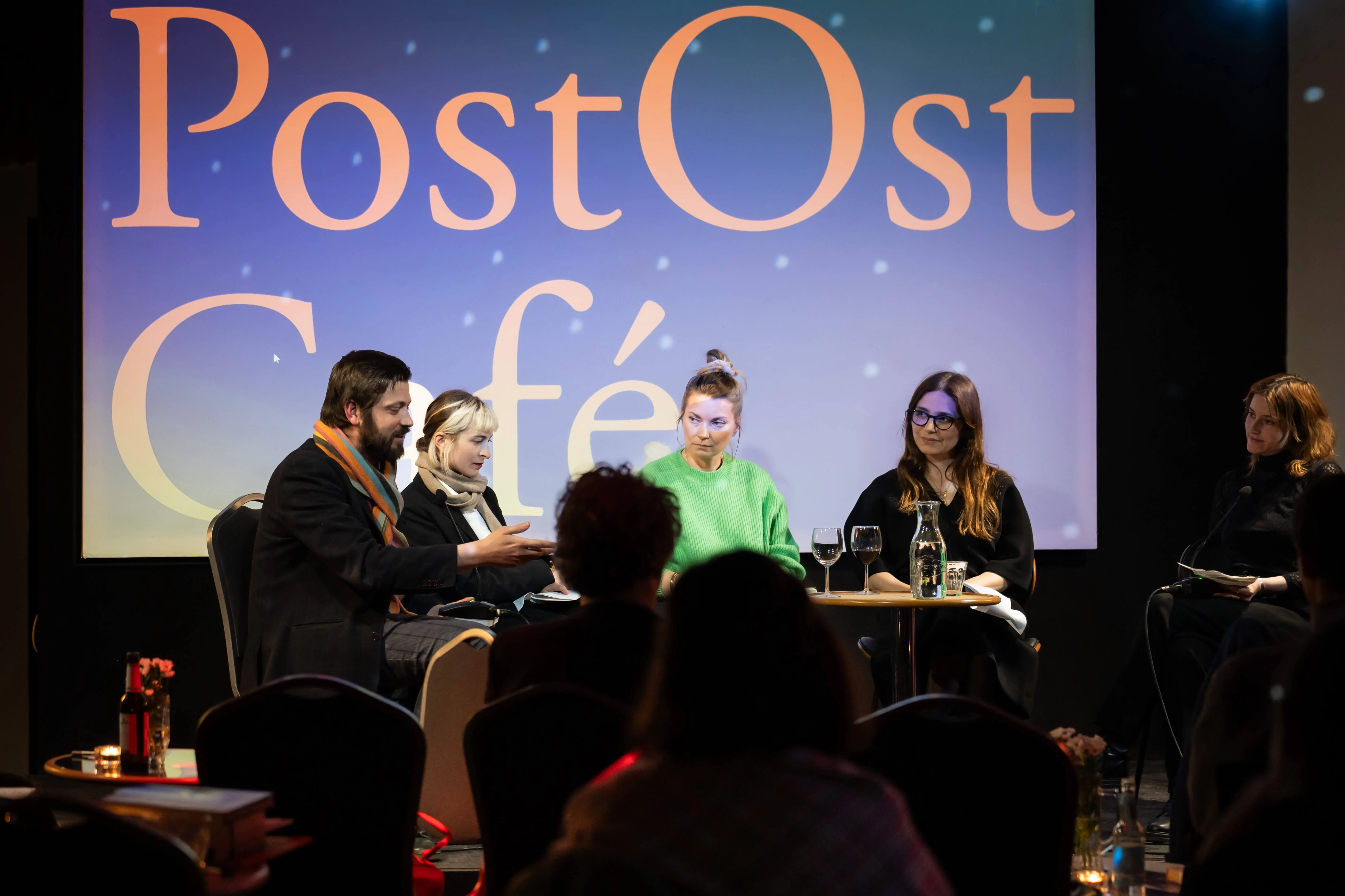 Fünf Menschen sitzen auf der Lettrétage-Bühne, im Hintergrund ist groß der Schriftzug "PostOstCafé" an die Leinwand projiziert.