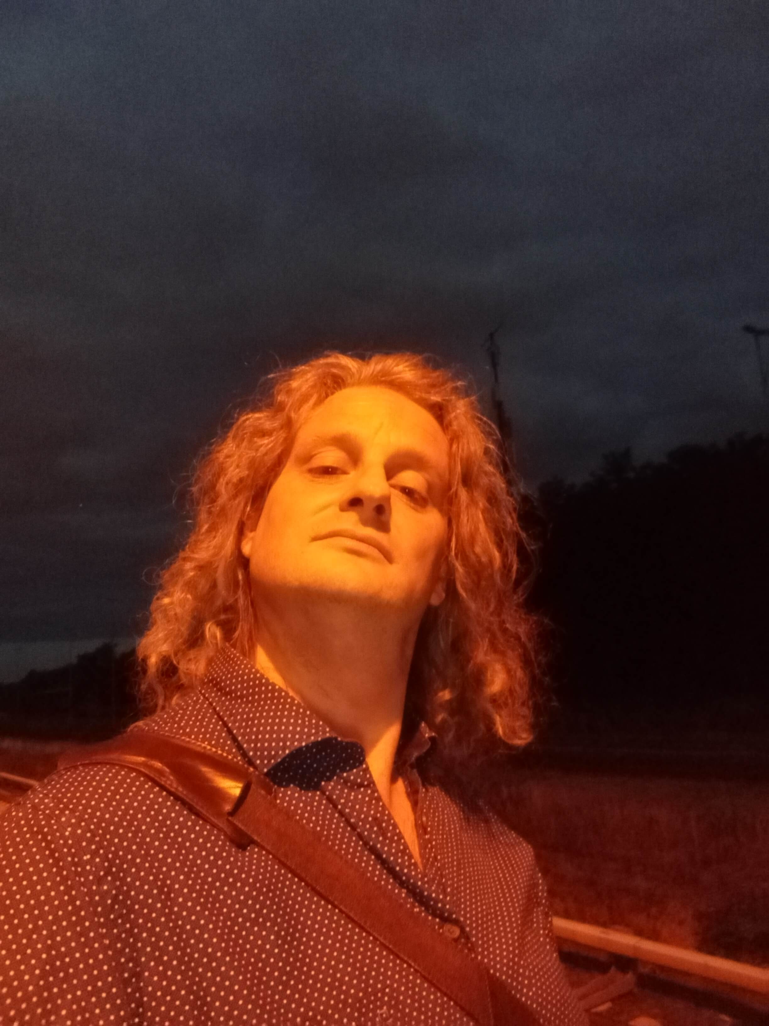 Porträtfoto von Fabian Herrmann, es ist dunkel, er wird von einer Straßenlaterne von oben beleuchtet, er schaut mit gehobenem Kopf in die Kamera und trägt ein dunkelblaues Hemd mit weißen Punkten, er hat schulterlange, gewellte Haare.