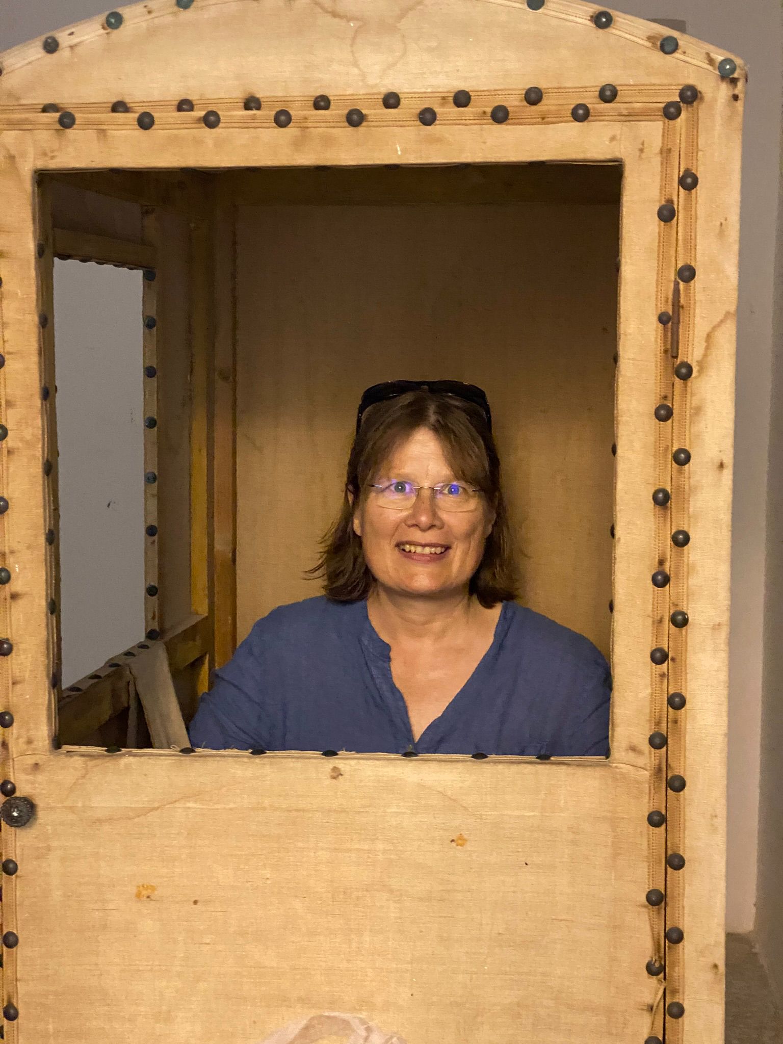 Porträtfoto von Carola Köhler, sie ist in einem kleinen Holzhäuschen und schaut lächelnd aus dem Fenster heraus.