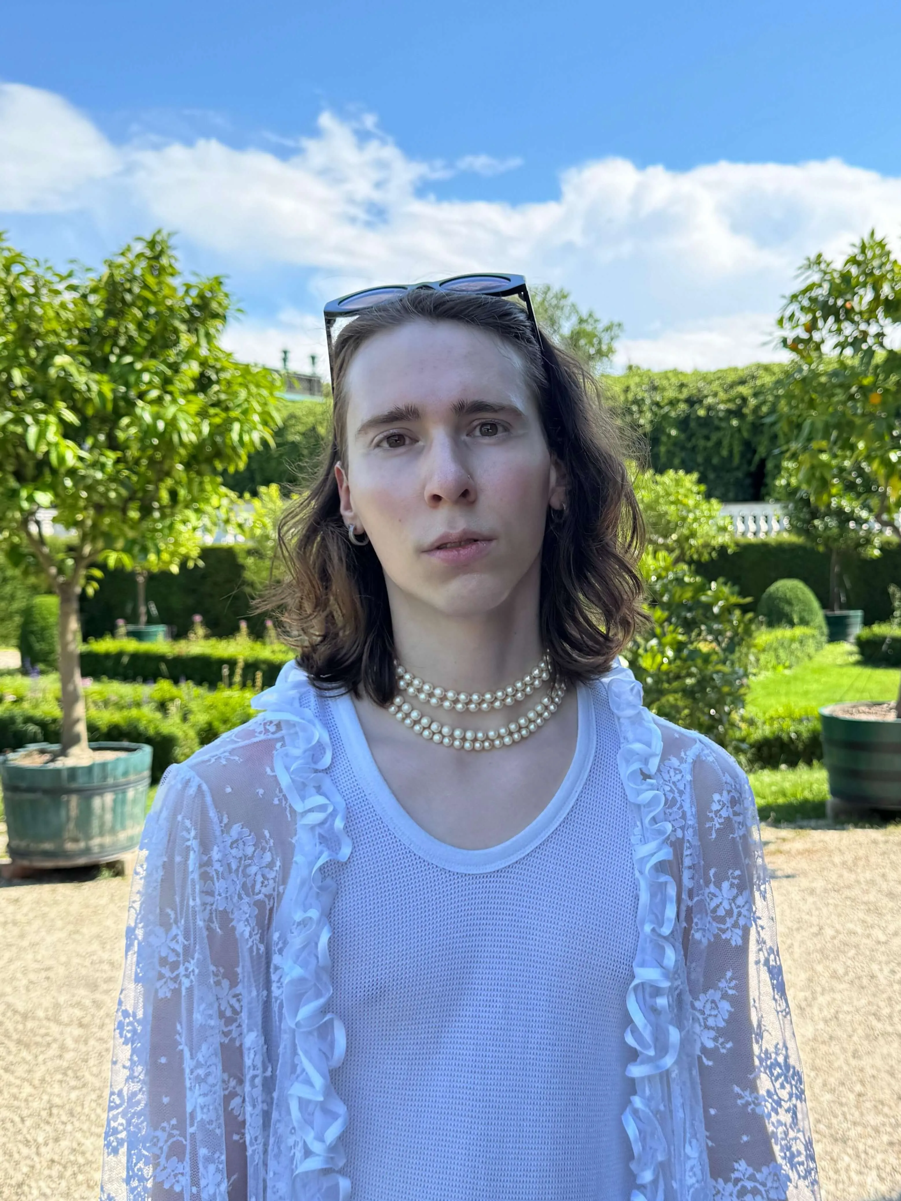 Porträtfoto von Toi Tautorus, sie schaut mit ernstem Blick in die Kamera, trägt eine weiße Spitzenbluse, eine Perlenkette und eine Sonnenbrille auf dem Kopf. Im Hintergrund: Blauer Himmel und ein leuchtend grüner Garten.