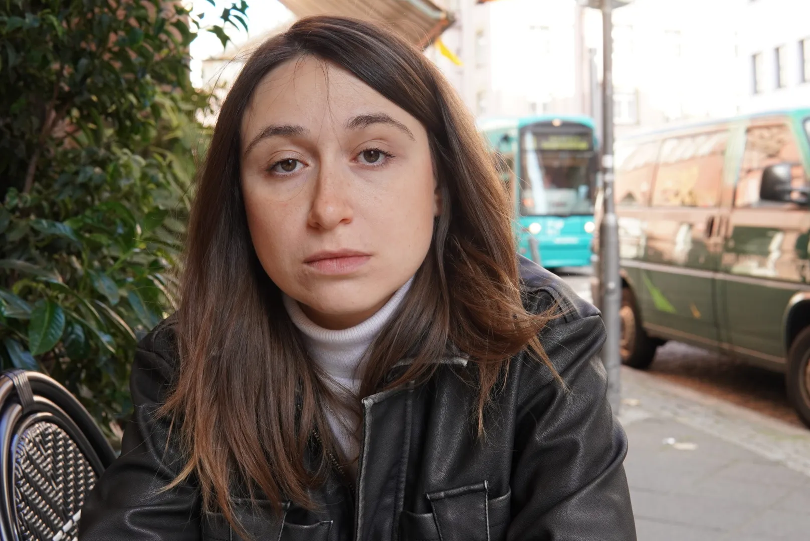 Porträtfoto von Maria Orlovskaya, sie sitzt an einem Tisch, hinter ihr fahren Autos auf einer Straße. Sie blickt ernst in die Kamera und trägt eine schwarze Lederjacke.