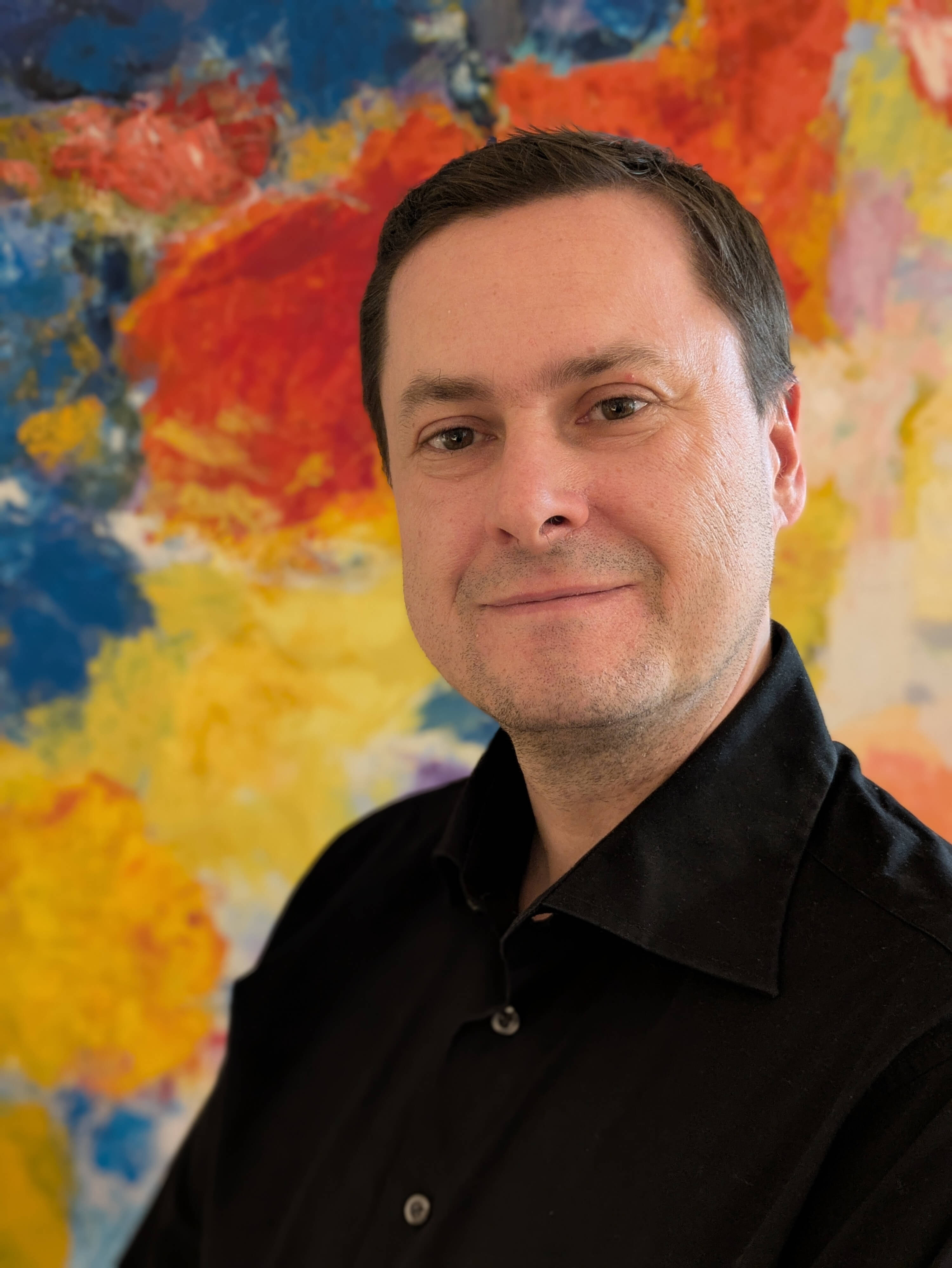 Porträtfoto von Tim Slagman, er lächelt in die Kamera, trägt ein schwarzes Hemd, im Hintergrund ist eine bunt bemalte Leinwand.