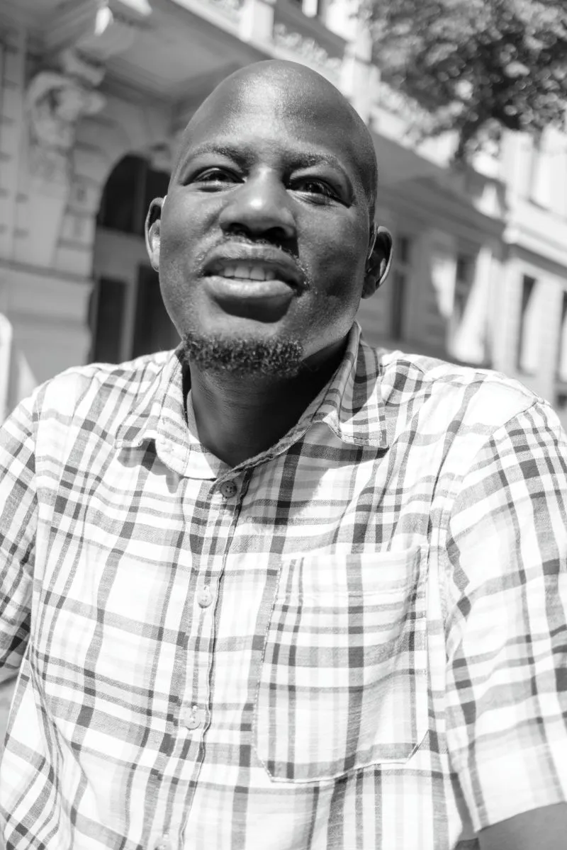 Schwarzweiß Portätfoto von Joe Otim Dramiga, er lächelt in die Kamera, trägt ein kariertes Hemd, im Hintergrund eine von der Sonne erleuchtete Hausfassade