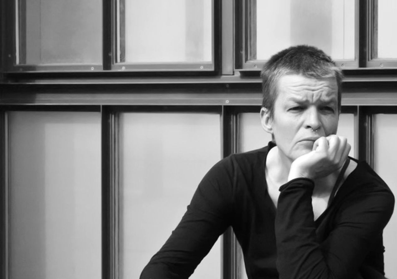 Schwarzweiß Porträtfoto von Susan Madsen. Sie hat ihren Kopf in ihre Hand gestützt und schaut ernst an der Kamera vorbei in die Ferne. Im Hintergrund sind symmetrische Ränder von bedeckten Fenstern.