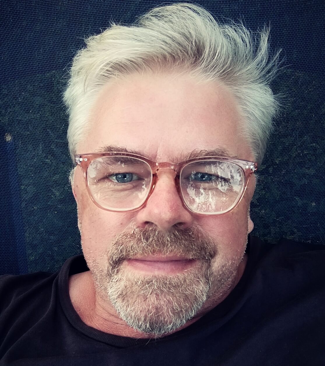 Selfie von Peter Tuschner. Er liegt auf einer Liege und blickt in die Kamera mit einem leichten Lächeln. Er trägt eine Brille, in der sich die Blätter einer Baumes spiegeln.
