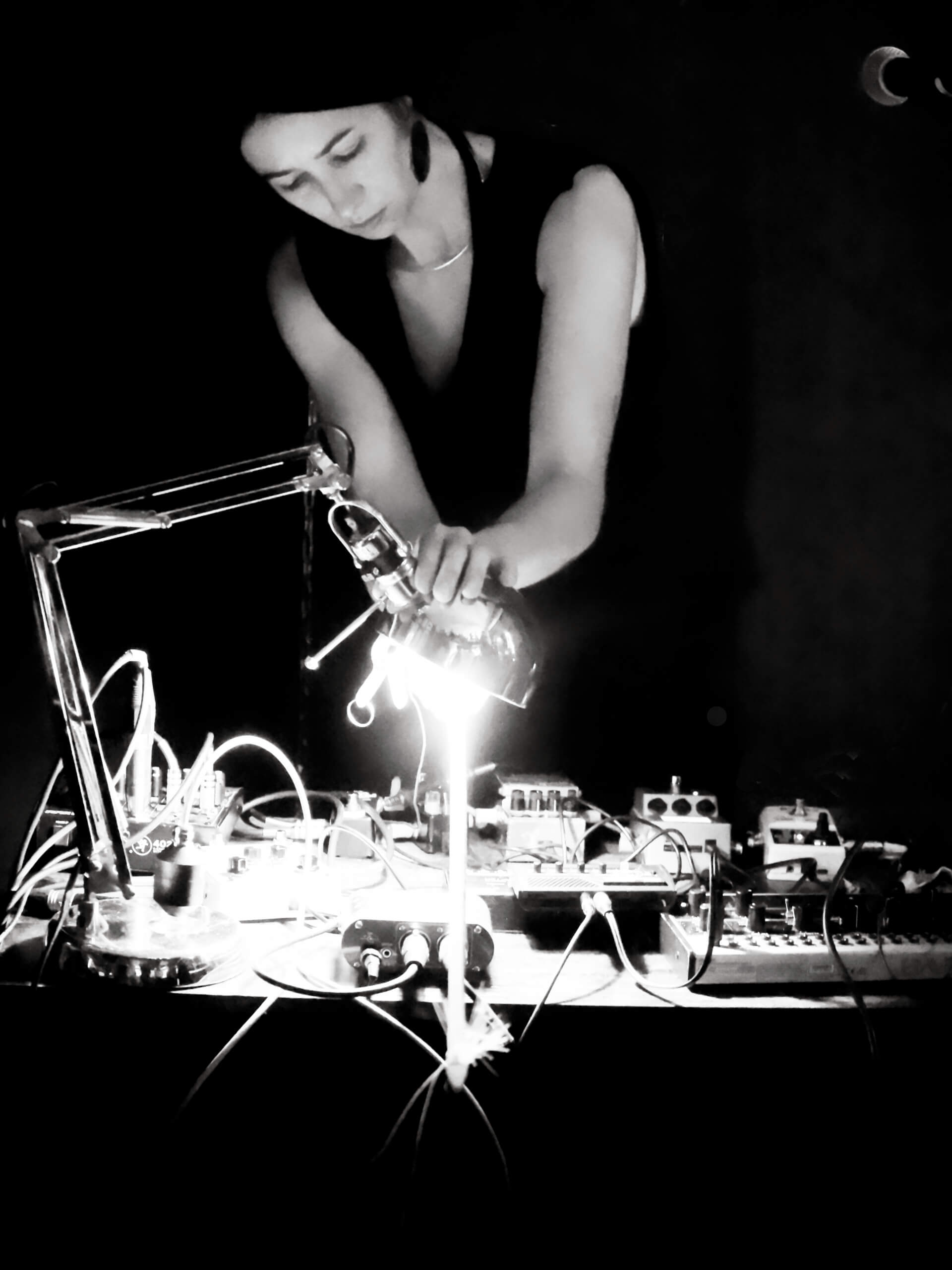 Schwarzweiß Foto von Cate Hops. Sie steht an einem Mischpult. Eine Schreibtischlampe projiziert einen hellen Lichtstrahl auf den Tisch, auf dem zahlreiche Geräte mit vielen Schaltern und Knöpfen stehen.