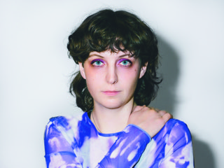 Porträtfoto von Fiona Sironic. Sie schaut ernst in die Kamera, der Hintergrund ist eine weiße Wand. Sie trägt ein blaues Batik Oberteil mit langen Ärmeln. Ihr Augen sind bunt geschminkt.