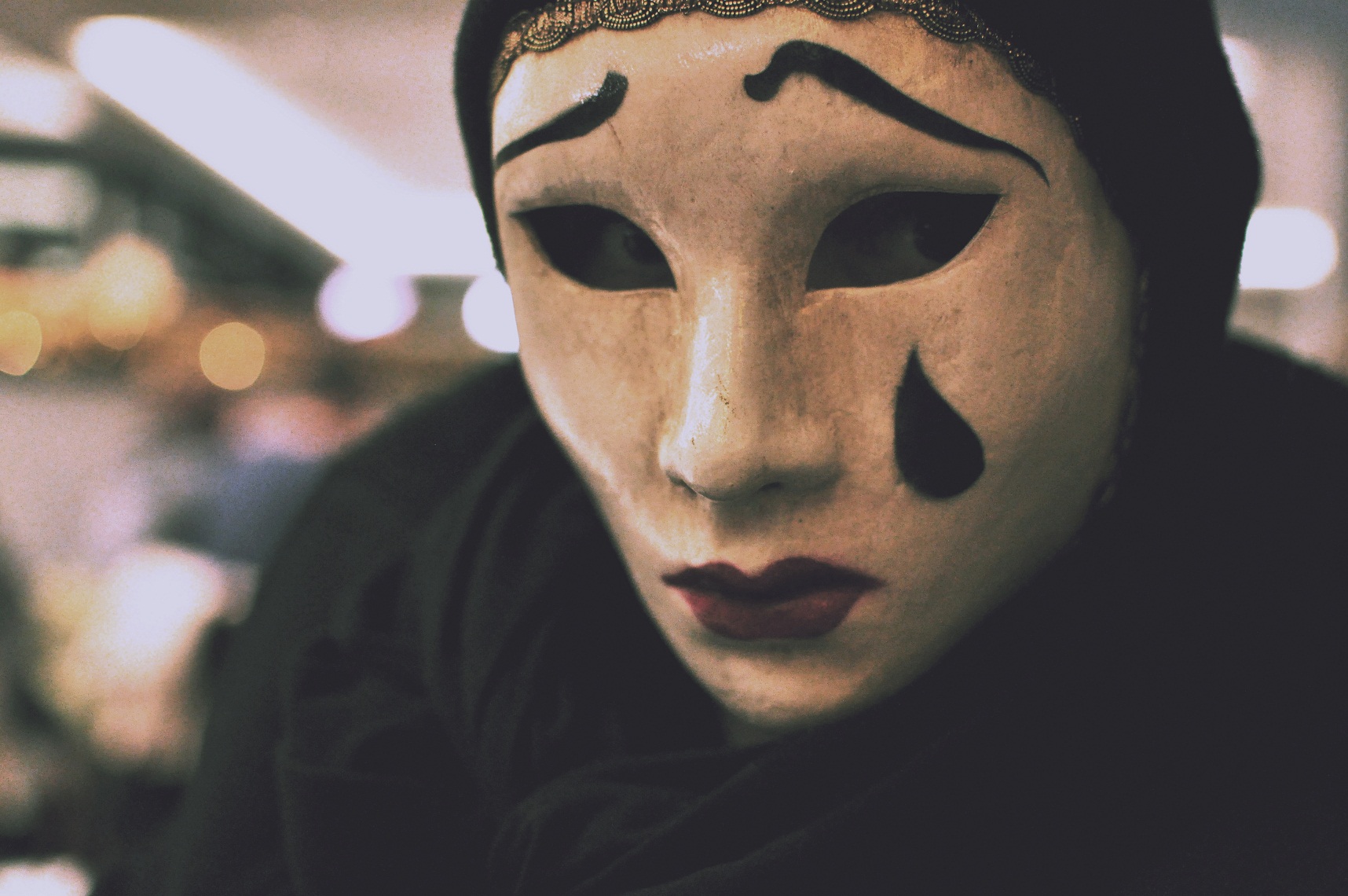 Porträtfoto von Sofie Lichtenstein, die eine Theatermaske trägt. Diese ist weiß, hat rote Lippen, schwarze, geschwungende Augenbrauen und eine schwarze Träne läuft über die linke Wange.