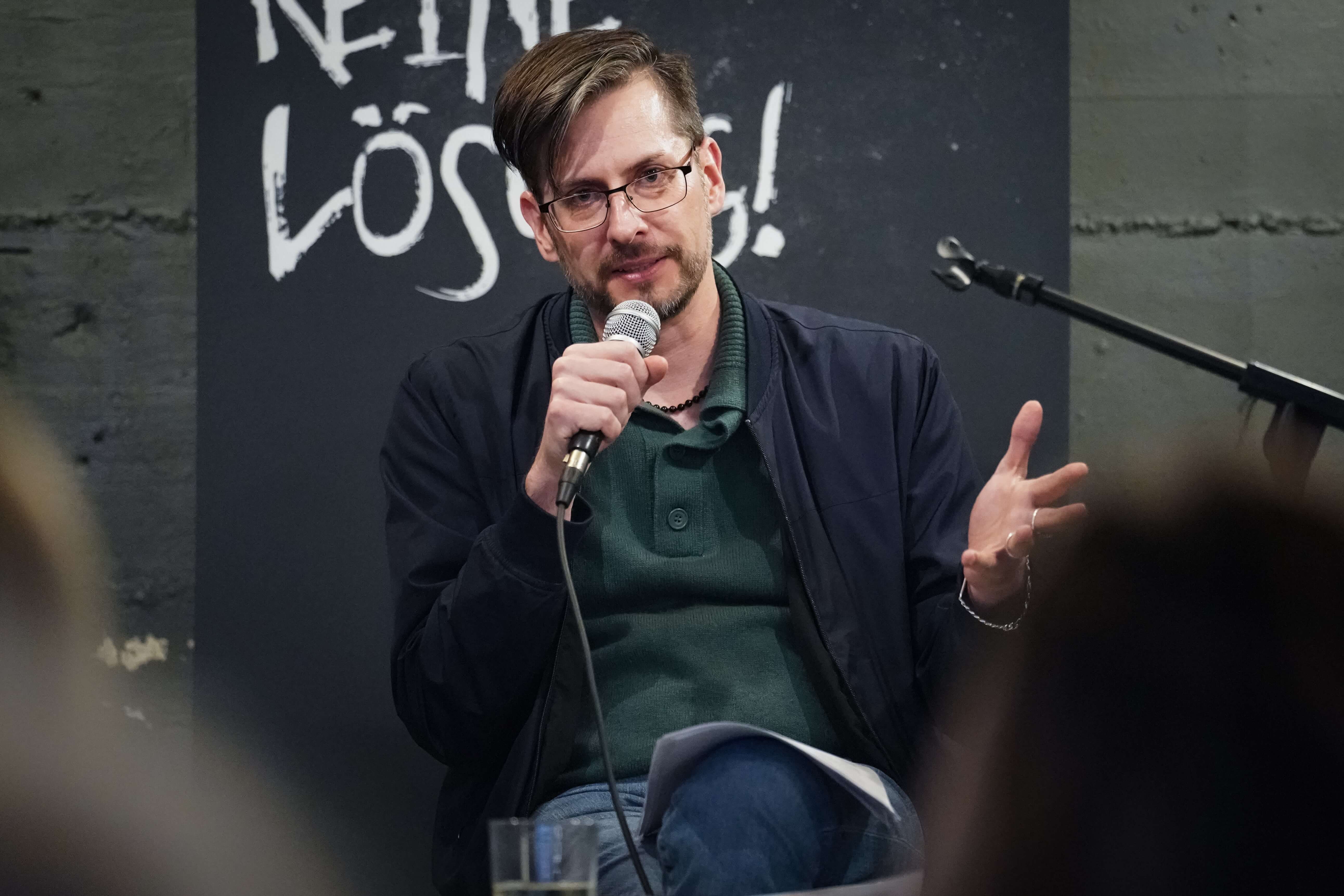 Foto von Alexander Graeff während einer Lesung. Er hält ein Mikrofon in der Hand, auf seinem Schoss liegen Blätter und er spricht mit dem Publikum.