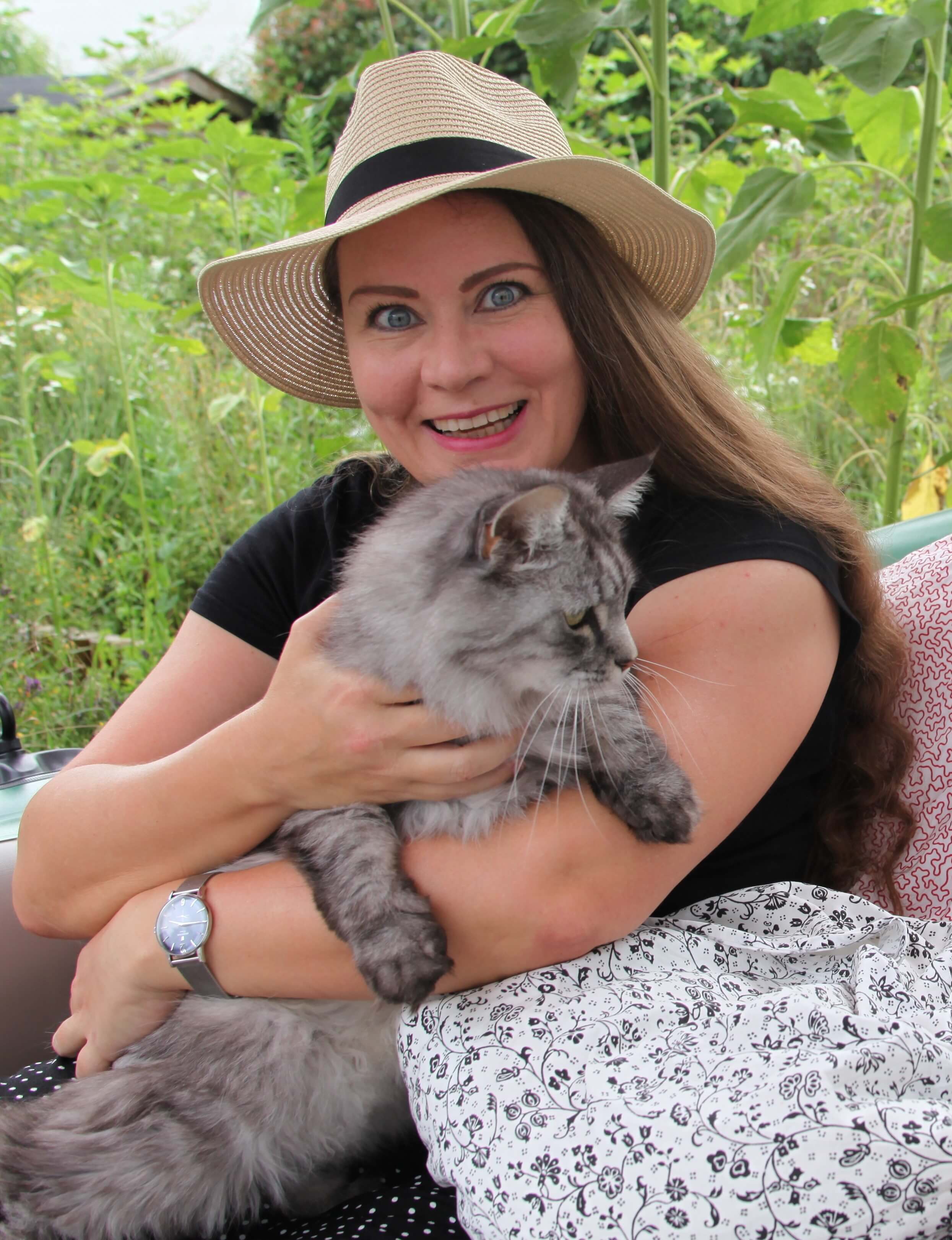 Foto von Tanja Gabriele Baudson. Sie sitzt in einem Garten, trägt einen Sonnenhut, lächelt in die Kamera und hält eine grau gestreifte Katze im Arm.