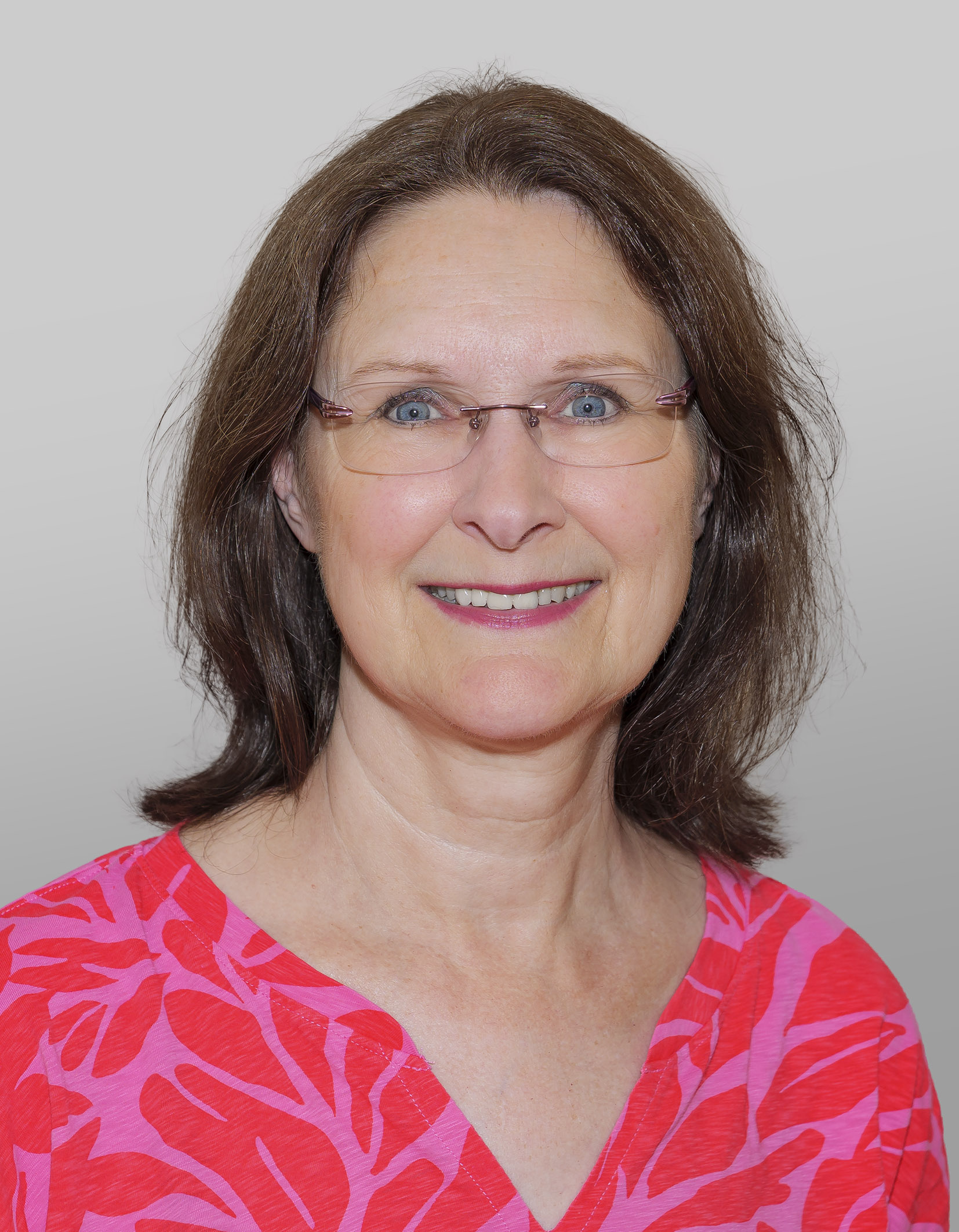 Porträtfoto von Kristen Boysen. Vor einem weißen Hintergrund lächelt sie in die Kamera. Sie trägt ihre schulterlangen, braunen Haare offen, sie trägt eine zarte Brille und eine pink-rote Bluse.